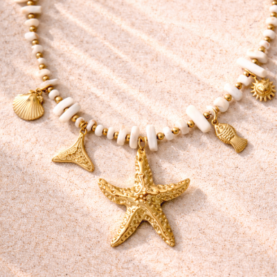 Colar branco e dourado com pendentes marinhos sobre areia clara