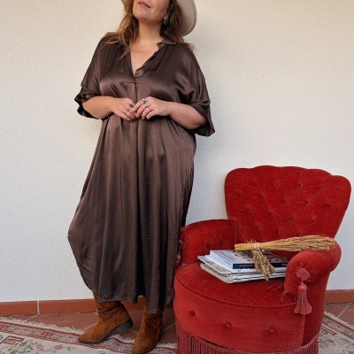 Mulher com vestido castanho acetinado e chapéu bege ao lado de poltrona vermelha e livros