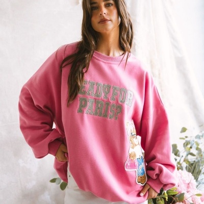 Mulher a vestir sweatshirt rosa com texto READY FOR PARIS? e patch de urso, calças brancas e fundo com plantas.