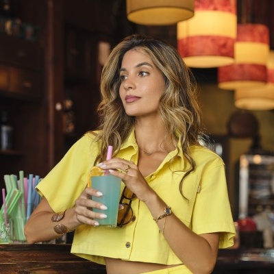 Mulher com roupa amarela segurando copo de bebida azul num bar