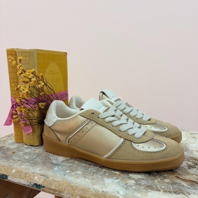 Ténis em camurça bege e pele metálica prateada com atacadores brancos em cima de uma mesa de madeira com livro e flores secas ao fundo