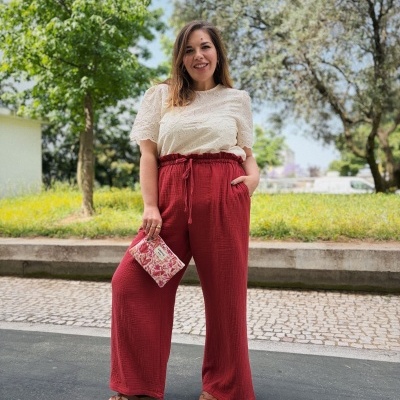 Mulher com blusa branca bordada, calças vermelhas largas, e mala estampada AMOUR ao ar livre