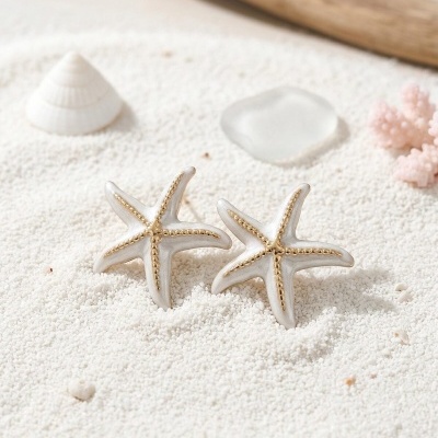 Brincos de estrela do mar branco com detalhes dourados na areia branca com concha, vidro, coral e madeira ao fundo