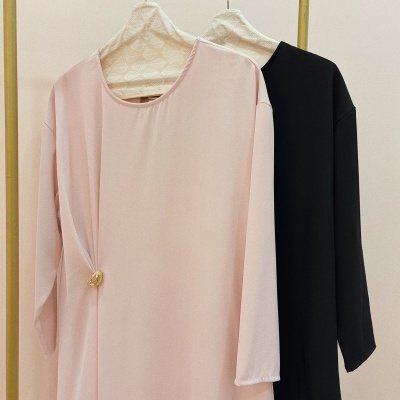 Duas túnicas, uma rosa clara com detalhe dourado e uma preta, penduradas em cabides com capa de folhas brancas num cabide de metal amarelo.