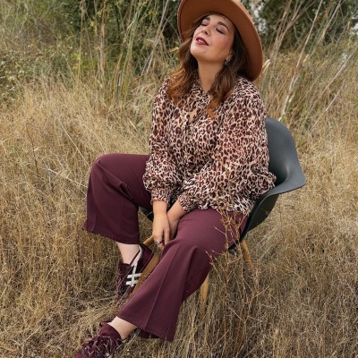 Pessoa sentada em cadeira ao ar livre com blusa padrão oncinha, calças bordô, boné castanho e sapatilhas bordô