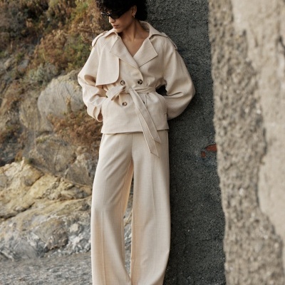 Mulher com fato bege e sapatos pretos encostada em parede exterior rochosa.