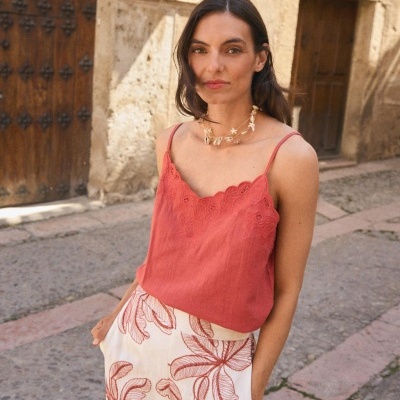 Mulher com blusa terracota e calças brancas com padrão de folhas em rua com porta de madeira