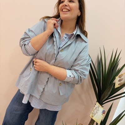 Mulher com casaco cinza claro, camisa xadrez azul e branco, jeans azuis, posando em ambiente interior perto de planta decorativa.