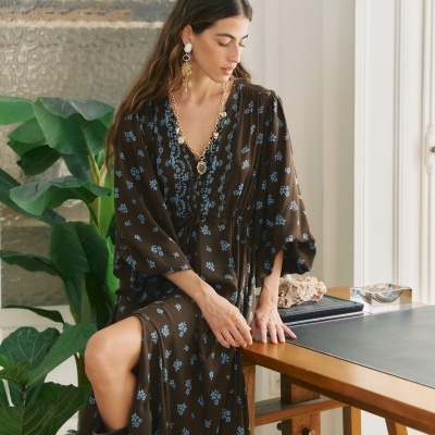 Mulher com vestido comprido castanho com flores azuis, colar dourado e brincos grandes, sentada junto a mesa de madeira.