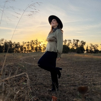 Mulher com vestido verde e chapéu preto em campo aberto ao pôr do sol.