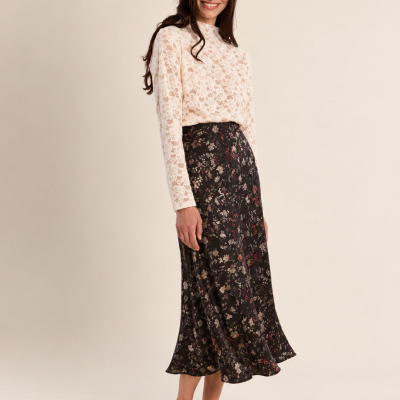 Modelo com blusa branca de renda e saia midi preta estampada em fundo neutro