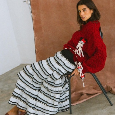 Mulher vestindo camisola vermelha de malha e saia longa branca com riscas pretas, sentada numa cadeira preta frente a fundo castanho.