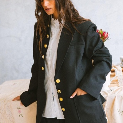 Mulher com casaco preto de botões dourados e camisa branca, em ambiente interior com mesa e flores