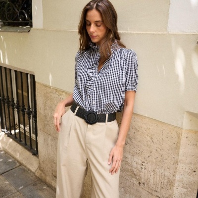Mulher veste blusa xadrez preta e branca com folhos e calças largas bege com cinto preto em rua com parede clara.