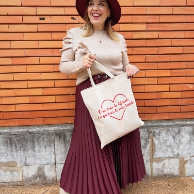 Mulher com chapéu vinho e tote bag bege com texto em fundo de tijolos laranja.