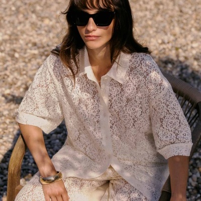Mulher de blusa e calças em renda bege com padrão floral, óculos escuros pretos, sentada em cadeira de madeira sobre chão de pedras