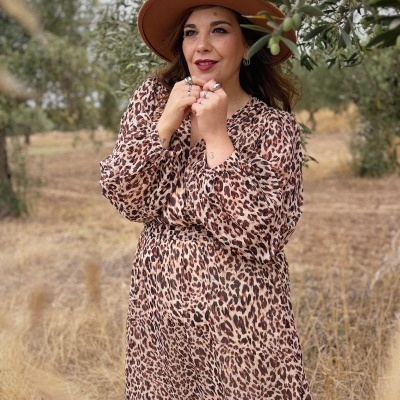 Mulher com vestido animal print e chapéu castanho num campo com árvores.