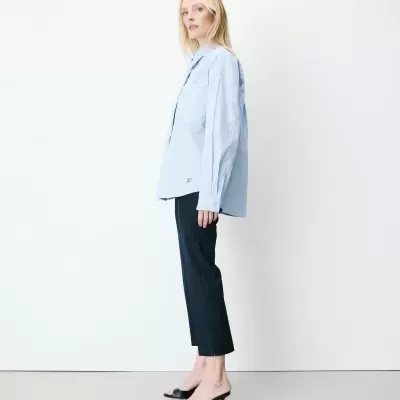 Mulher loira a usar camisa azul riscada e calças pretas com sapatos de tacão preto, em fundo branco