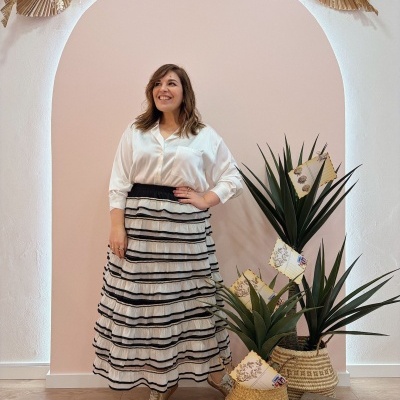 Mulher com camisa branca e saia longa folhada em ambiente decorado com plantas e arco rosa