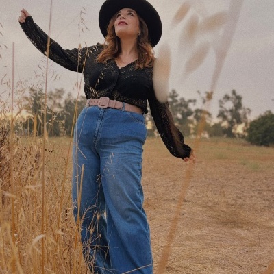 Mulher com blusa preta de renda, calças de ganga azul, cinto bege e chapéu preto em campo seco
