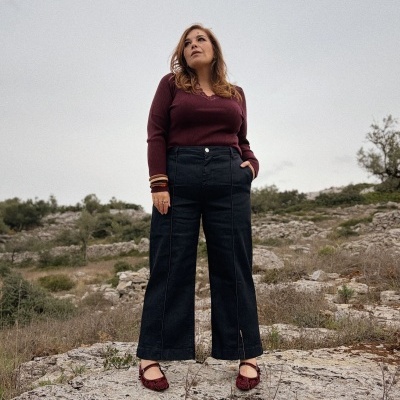 Mulher com camisola bordeaux, calças pretas largas e sapatilhas bordô numa paisagem rural rochosa.