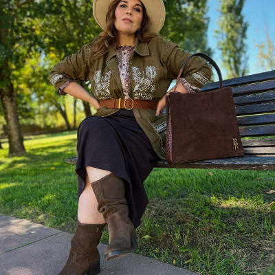 Mulher sentada no banco de parque com botas e acessórios castanhos e casaco verde bordado.