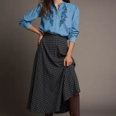 Mulher com camisa azul bordada, saia xadrez e botas castanhas.