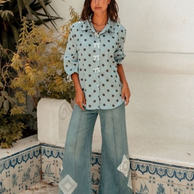 Mulher com camisa azul clara de bolinhas pretas e calças largas de ganga com remendos de renda, sapatos pretos, em ambiente exterior com azulejos e plantas