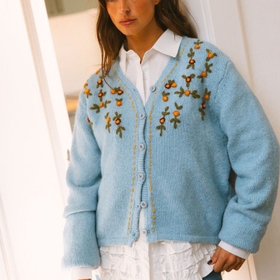 Mulher com cardigã azul claro bordado sobre camisa branca e calças jeans em interior.