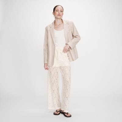 Mulher com blazer bege, top branco, calças rendadas brancas e sandálias pretas
