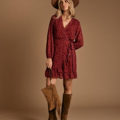 Mulher com vestido vermelho de padrão pequeno, botas e mala castanhas, chapéu castanho, fundo castanho.