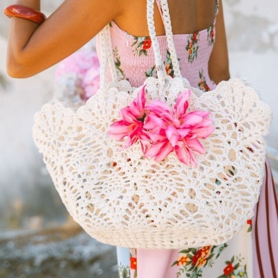 Mala de crochê branco com flores rosas e alças trançadas em ombro de mulher com vestido floral.