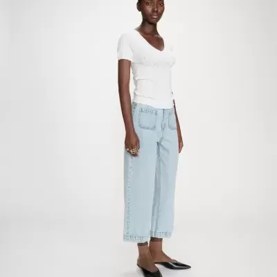 Mulher a usar camisola branca, calças de ganga azul claro, meias castanhas e sapatos pretos num fundo branco.