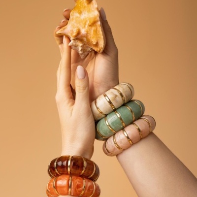 Mãos femininas com pulseiras grossas coloridas seguram uma concha amarela, fundo bege.