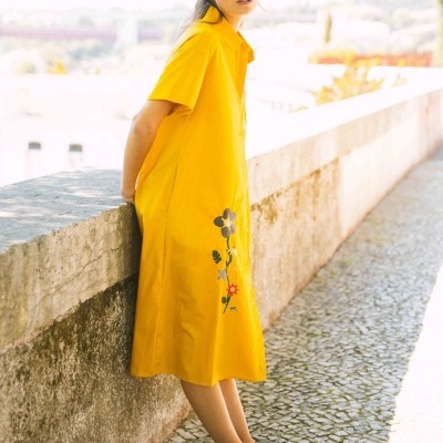 Vestido amarelo curto com bordado floral usado por modelo junto a muro de pedra em calçada de calhaus