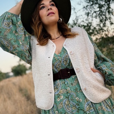 Mulher com vestido verde estampado, colete bege felpudo e chapéu verde-oliva ao ar livre