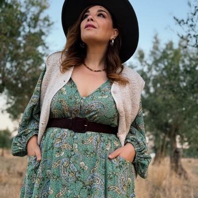 Mulher com vestido verde estampado, cinto castanho, colete bege felpudo e chapéu preto em ambiente exterior