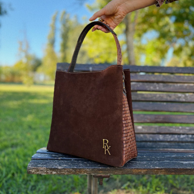 Bolsa de senhora castanha em camurça com padrão trançado e letras douradas RK num banco de madeira