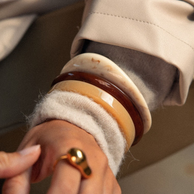 Pulso com três pulseiras castanhas e bege, camisola clara felpuda, e mão com anéis dourados e pedra roxa.