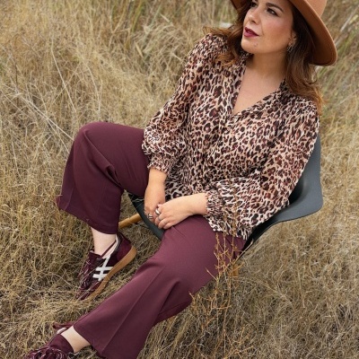 Mulher com blusa animal print, calças vinho, sapatilhas vinhas e chapéu castanho sentada na natureza