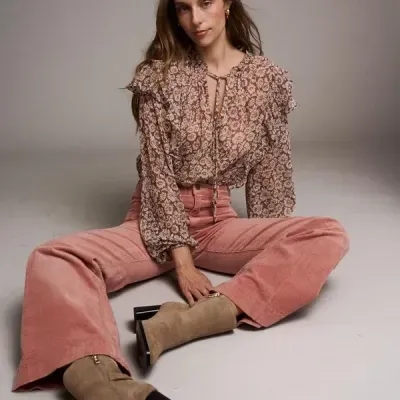 Mulher com blusa floral castanha, calças rosa e botas bege sentada no chão com fundo cinza