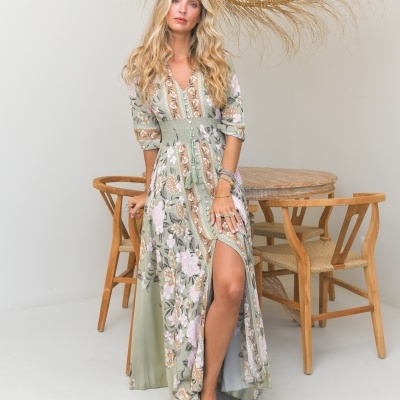 Vestido longo verde floral com fenda e botões, modelo loira posando em ambiente minimalista.