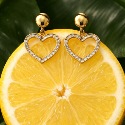 Brincos dourados em forma de coração com pedras sobre metade de limão e folhas verdes ao fundo
