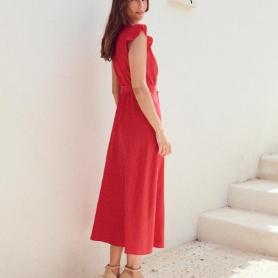 Mulher com vestido comprido vermelho e sandálias de cunha em ambiente com parede branca e escadas de pedra.