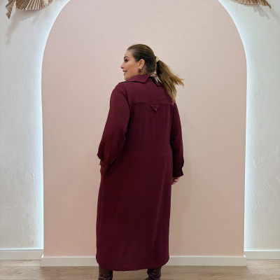 Mulher com vestido vinho e botas castanhas de costas em fundo de arco e plantas secas.