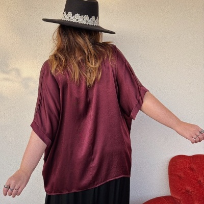 Pessoa com chapéu preto decorado e camisa vinho acetinada vista de costas junto a cadeira vermelha.