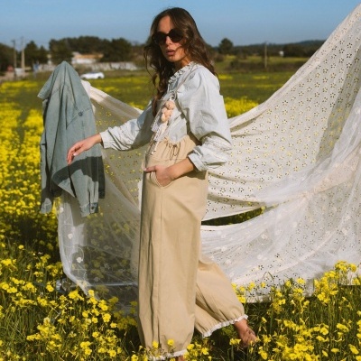 Mulher com blusa bege clara, calças largas bege, óculos de sol em campo de flores amarelas com tecido rendado branco ao fundo