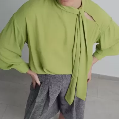 Blusa verde com laço no decote e calças cinzentas largas em ambiente interior.