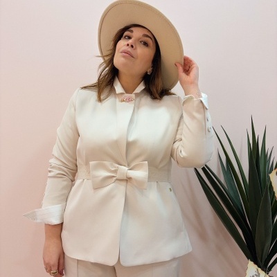 Mulher vestindo conjunto bege com blazer de laço e chapéu bege ao lado de planta verde