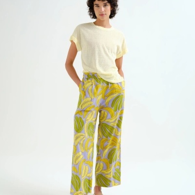 Mulher com t-shirt amarela clara e calças largas com padrão de bananas amarelas e verdes, calçado bege em fundo branco